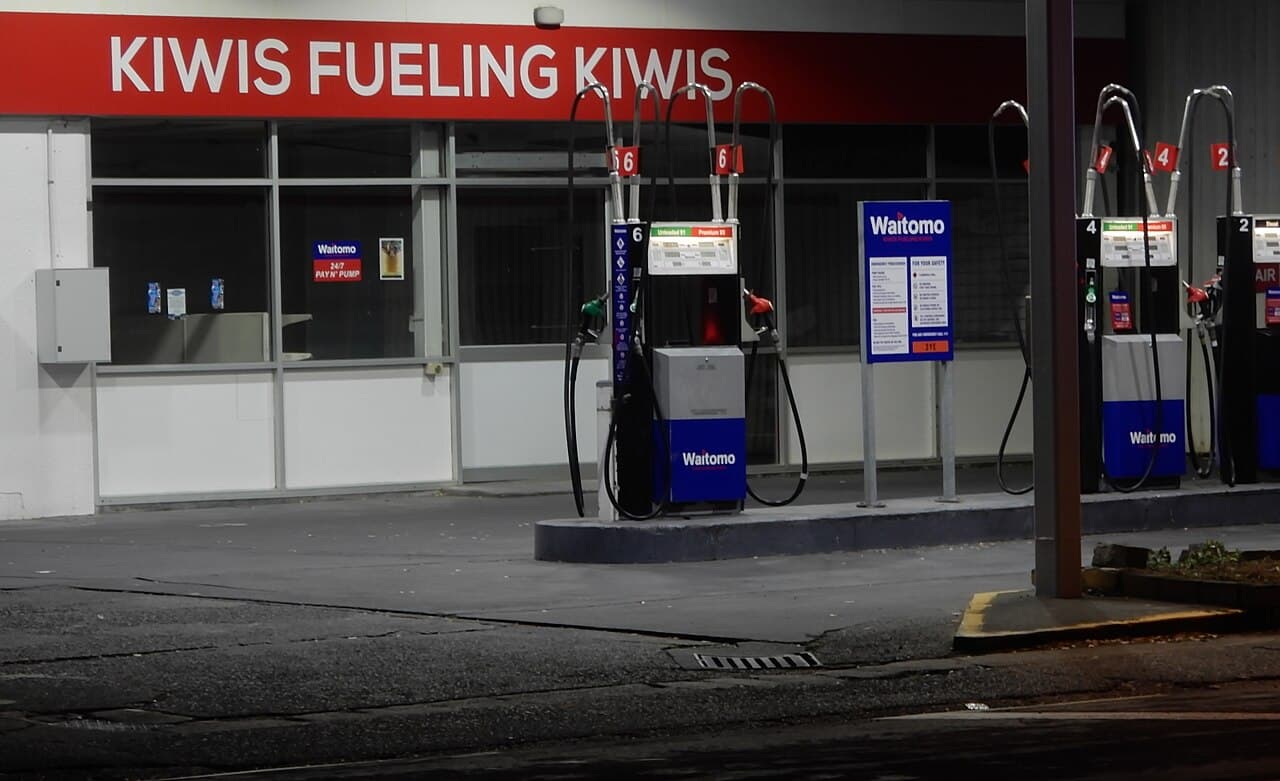 A New Zealand service station forecourt at night, unleaded and premium pumps under a sign reading Kiwis Fueling Kiwis