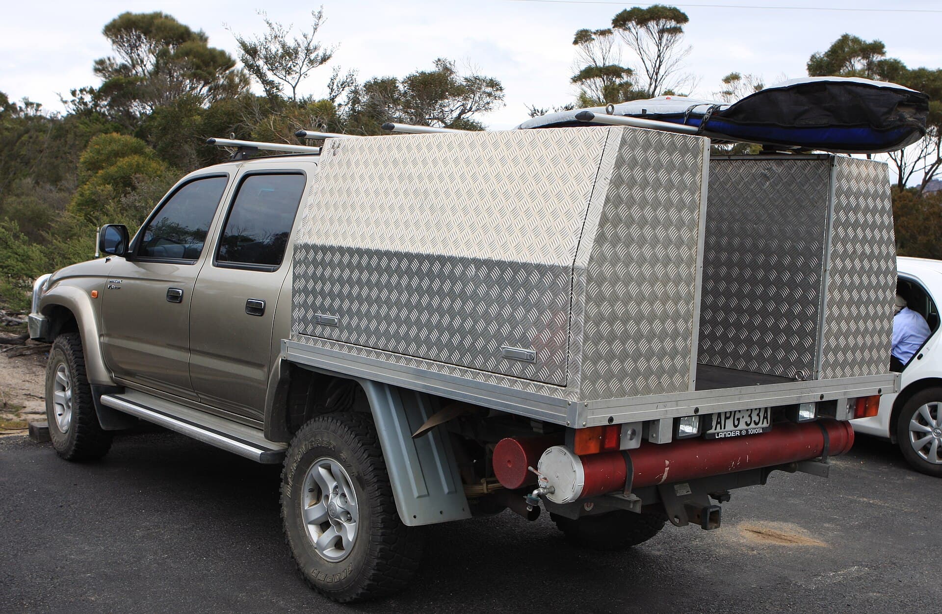 A dual-cab Toyota HiLux fitted with an aluminium tray, a toolbox canopy, a roof rack and gas bottles, parked on a bush track